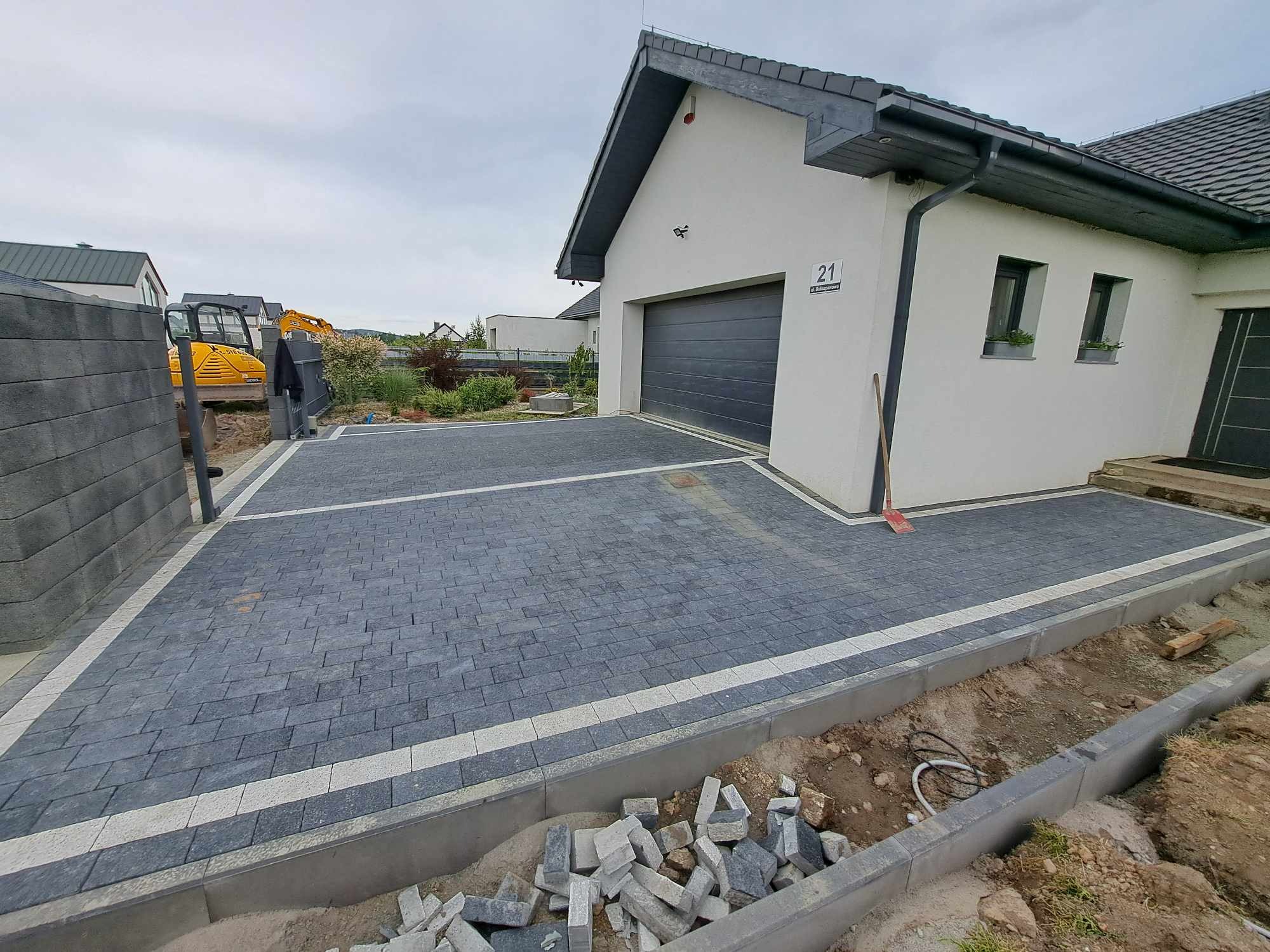 Nowa, szara kostka brukowa ułożona na podjeździe przed nowoczesnym domem z szarą bramą garażową i numerem 21, widoczny fragment wykopu i maszyna budowlana w tle.