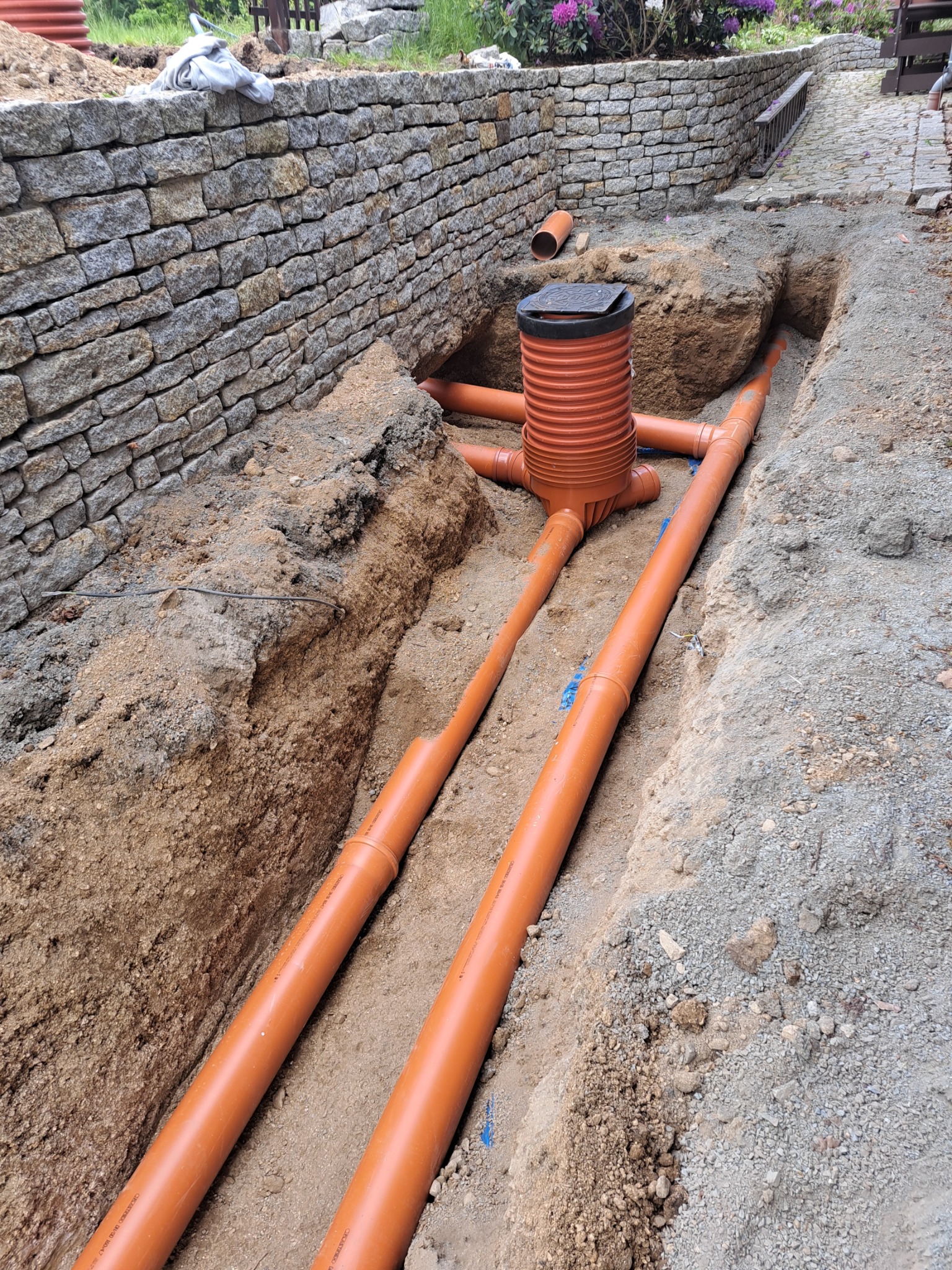 Pomarańczowe rury kanalizacyjne ułożone w wykopie, widoczna studzienka rewizyjna z pokrywą, w tle kamienny murek oporowy i fragment chodnika.
