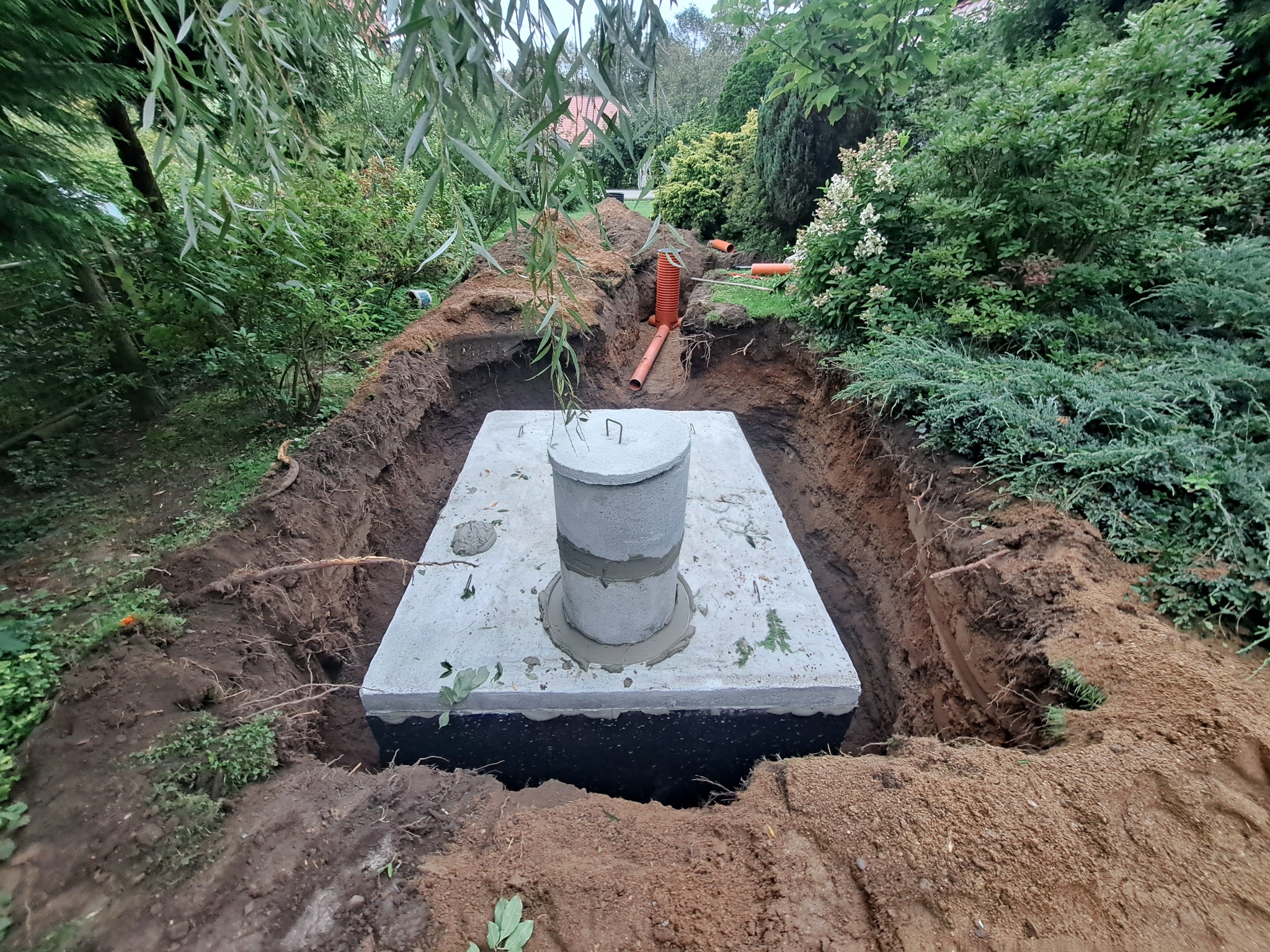 Betonowy zbiornik z pokrywą, umieszczony w wykopie ziemnym, otoczony zielenią i pomarańczowymi rurami drenażowymi.