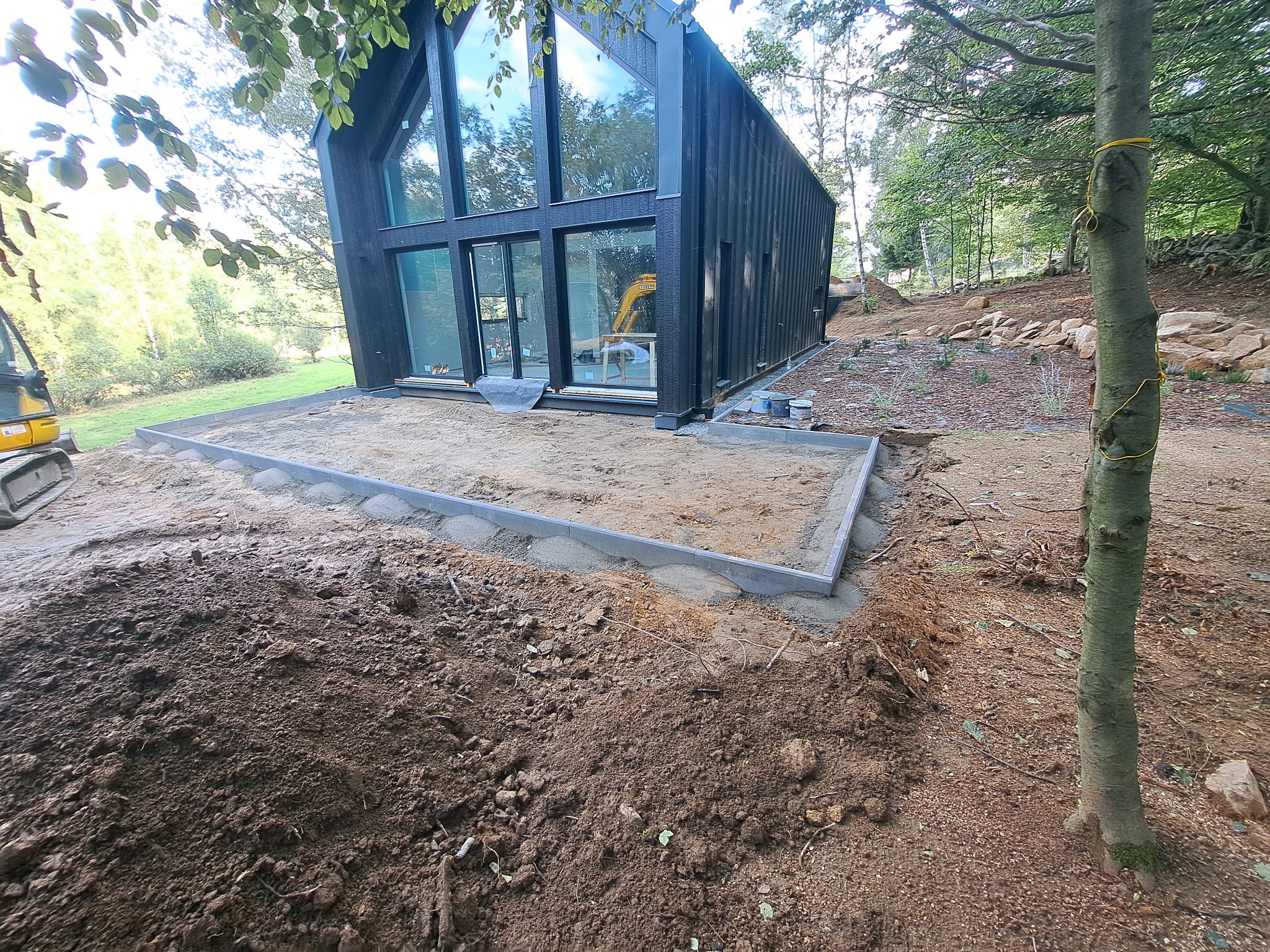 Przygotowanie podłoża z piasku i obrzeży betonowych pod taras przy nowoczesnym, czarnym domu z dużymi oknami; w tle widoczny minikoparka i drzewa.