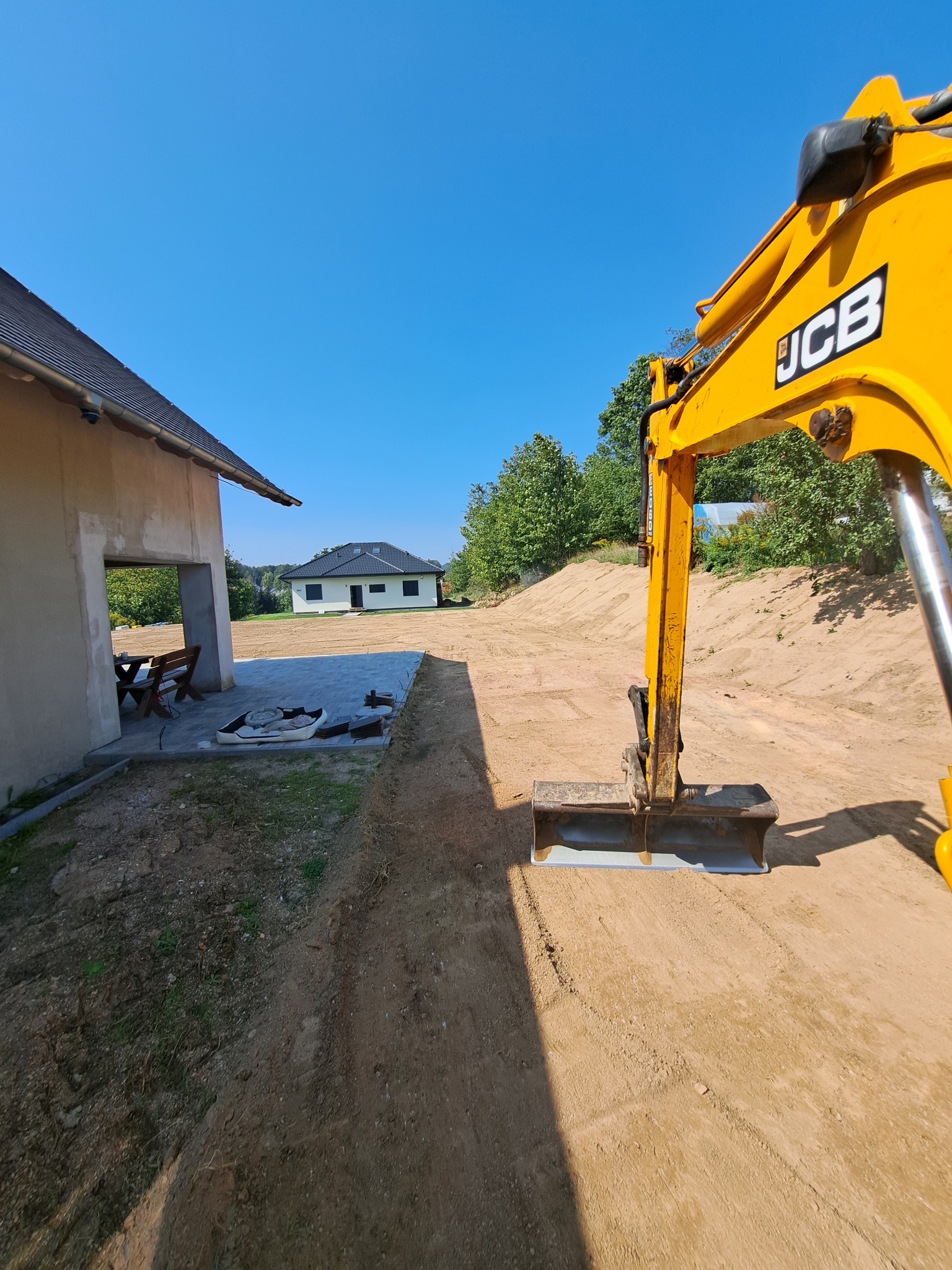 Żółta koparka JCB przygotowuje teren pod budowę domu, widoczny świeżo wyrównany grunt i częściowo ułożona kostka brukowa na podjeździe.