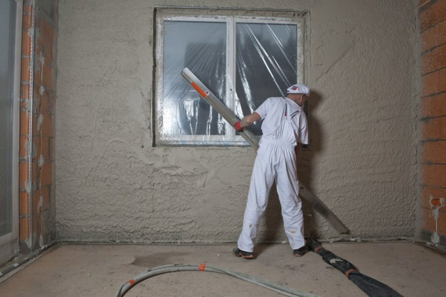 Aplikacja tynku maszynowego na ścianę wewnętrzną, pracownik w białym kombinezonie wyrównuje powierzchnię łatą, okno zabezpieczone folią.