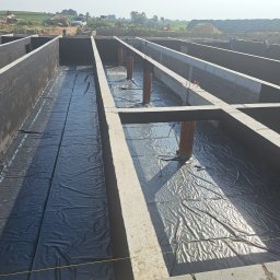 WELDIS SPÓŁKA Z OGRANICZONĄ ODPOWIEDZIALNOŚCIĄ - Zalane fundamenty z izolacją przeciwwilgociową z folii, widoczne wyprowadzenia rur, ściany fundamentowe z bloczków betonowych, teren budowy w tle.