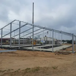 Konstrukcja stalowa hali w trakcie montażu, widoczne fundamenty i podłoże z piasku, w tle budynek gospodarczy i dźwig.