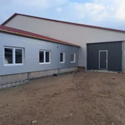 Nowoczesny budynek przemysłowy z szarymi panelami, czerwonym dachem i dużą, ciemną bramą garażową, widziany z zewnątrz na terenie budowy z błotnistym podłożem i rozłożoną drabiną.