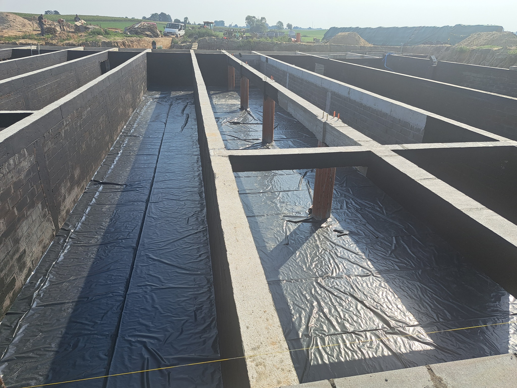 Zalane fundamenty z izolacją przeciwwilgociową z folii, widoczne wyprowadzenia rur, ściany fundamentowe z bloczków betonowych, teren budowy w tle.