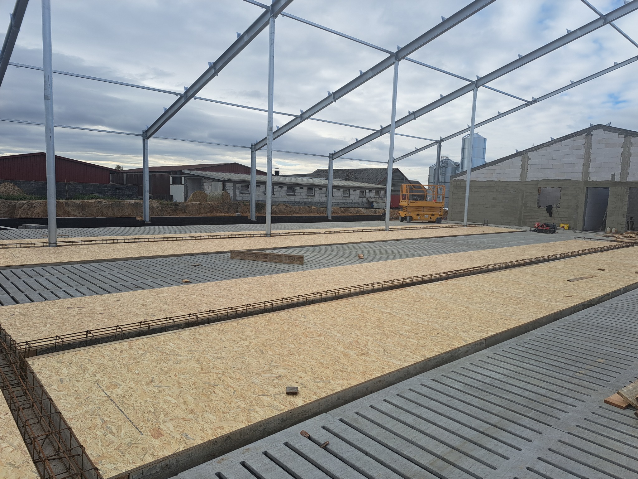 Wznoszenie konstrukcji budynku gospodarczego: stalowy szkielet, prefabrykowane płyty stropowe zbrojone stalą i drewniane szalunki na fundamencie.