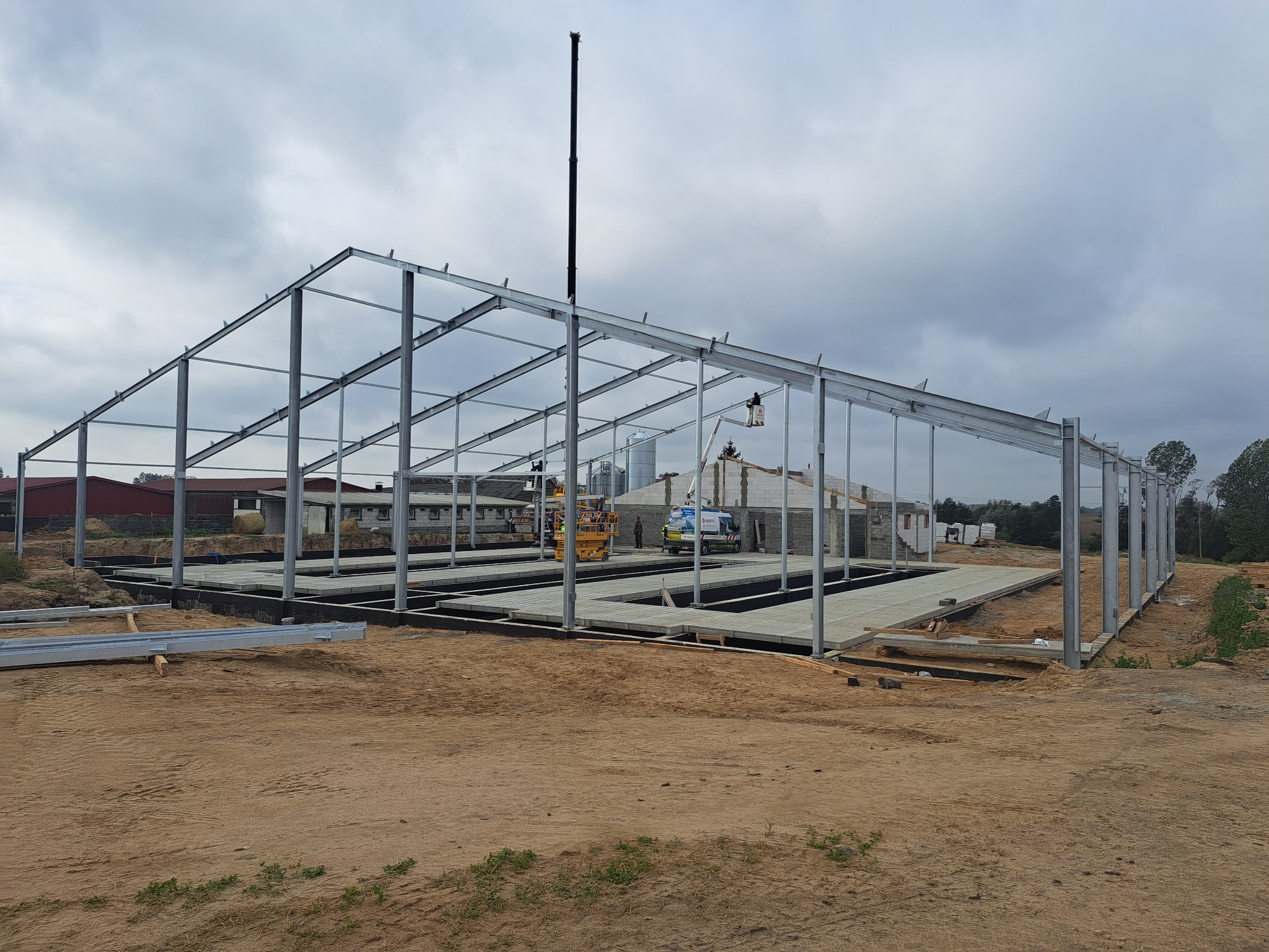Konstrukcja stalowa hali w trakcie montażu, widoczne fundamenty i podłoże z piasku, w tle budynek gospodarczy i dźwig.