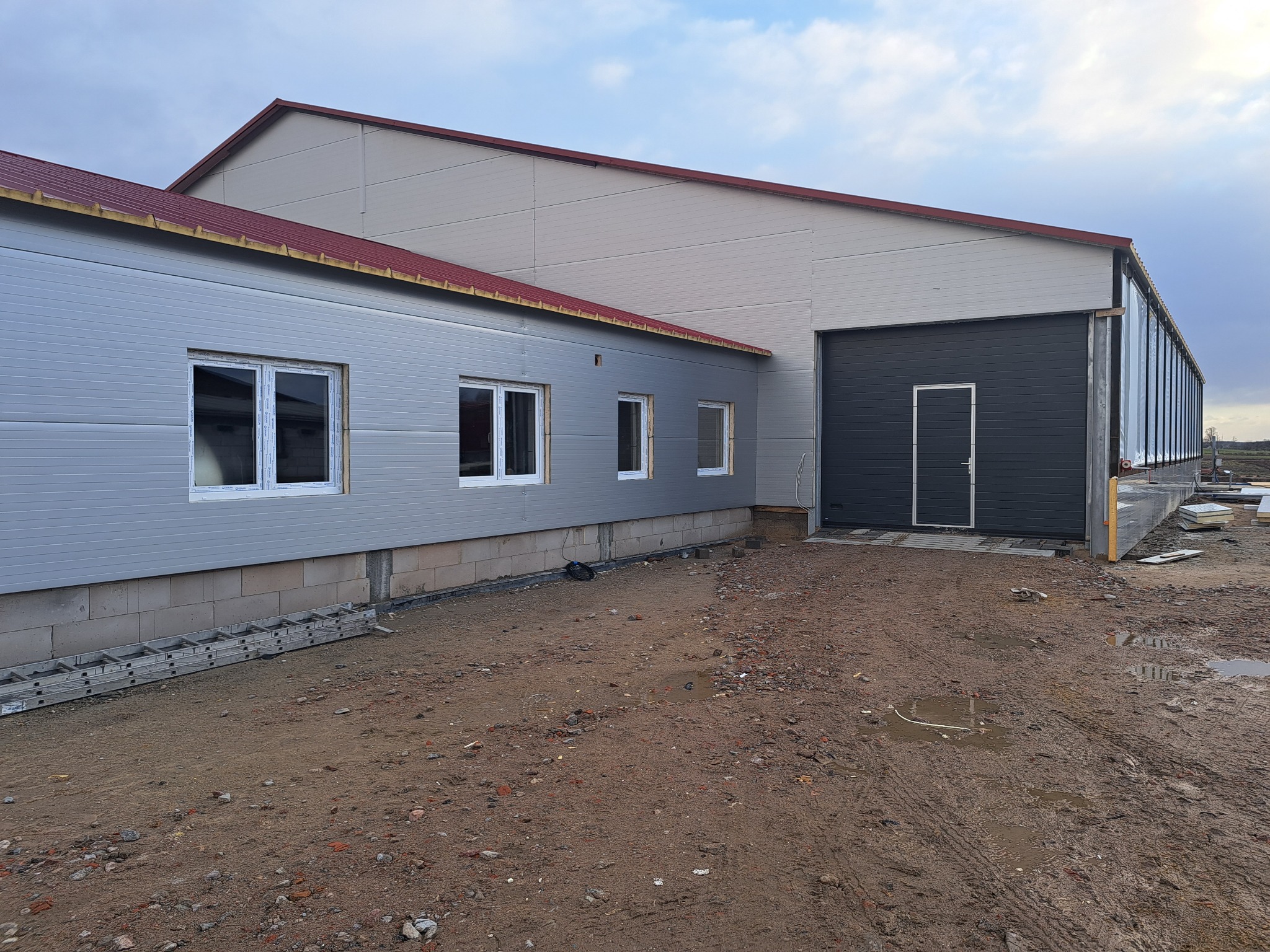 Nowoczesny budynek przemysłowy z szarymi panelami, czerwonym dachem i dużą, ciemną bramą garażową, widziany z zewnątrz na terenie budowy z błotnistym podłożem i rozłożoną drabiną.