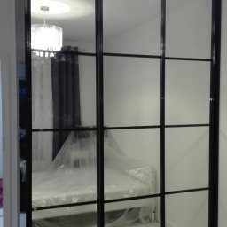 Meble w Twojej okolicy - Czarna szafa przesuwna z lustrzanymi drzwiami, widok na łóżko przykryte folią ochronną i lampę sufitową