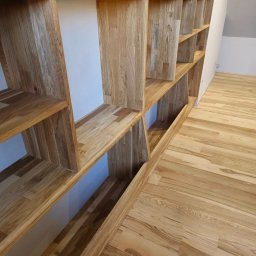 Meble w Twojej okolicy - Drewniana, modułowa półka na książki lub dekoracje, wkomponowana we wnękę, z widoczną strukturą drewna, dopasowana kolorystycznie do drewnianej podłogi.