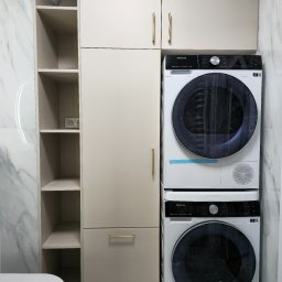 Meble w Twojej okolicy - Biała pralka i suszarka Hisense ustawione jedna na drugiej, obok wysoka zabudowa meblowa z półkami i szafkami w kolorze beżowym, łazienka wykończona marmurowymi płytkami.