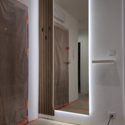 Meble w Twojej okolicy - Lustro z podświetleniem LED wkomponowane w drewniane lamele na ścianie, widoczne drzwi zabezpieczone folią malarską, gniazdko elektryczne z podłączonym kablem.
