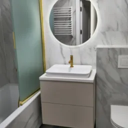 Nowoczesna łazienka z umywalką nablatową, szafką z szufladami w kolorze beżowym, baterią w kolorze złotym i okrągłym lustrem z podświetleniem LED na tle marmurowych płytek ściennych.