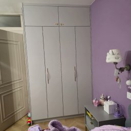 Meble w Twojej okolicy - Szara szafa na wymiar z złotymi uchwytami w pokoju dziecka z fioletowymi ścianami. Na podłodze leżą zabawki i ubrania, co dodaje wnętrzu przytulności.