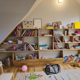 Meble w Twojej okolicy - Drewniana, nieregularna biblioteczka pod skosem na poddaszu, wypełniona książkami, zabawkami i dekoracjami w pokoju dziecka. Widoczny fragment dywanu i biurka.