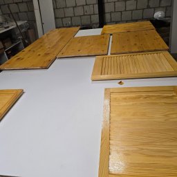 Meble w Twojej okolicy - Lakierowane, drewniane fronty meblowe (w tym żaluzjowe) leżą na białym stole w warsztacie. Widoczne słoje drewna i połysk lakieru. Część elementów poza kadrem.