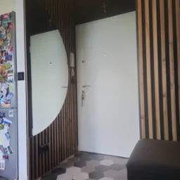 Nowoczesna aranżacja przedpokoju z lamelami drewnianymi, owalnym lustrem i heksagonalną podłogą. Nad drzwiami widoczny krzyż. Lodówka z magnesami po lewej.