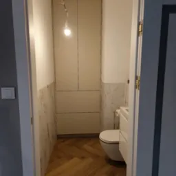 Nowoczesna szafa wnękowa w łazience w Ulan-Majorat. Białe fronty, marmurowe wykończenie ścian, parkiet w jodełkę. Widok z korytarza przez otwarte drzwi.