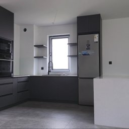 Meble w Twojej okolicy - Nowoczesna, minimalistyczna kuchnia z grafitowymi frontami, wyspą z białym blatem i sprzętem AGD w zabudowie. Widoczne półki wiszące i szara podłoga.