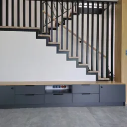 Nowoczesne schody z czarną balustradą i drewnianymi stopniami, zintegrowane z szarą szafką na ścianie. Widoczne gniazdka elektryczne. Minimalistyczny design.