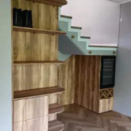 Drewniana zabudowa pod schodami z półkami, szafkami i chłodziarką do wina. Parkiet ułożony w jodełkę. Nowoczesne schody ze szklaną balustradą.