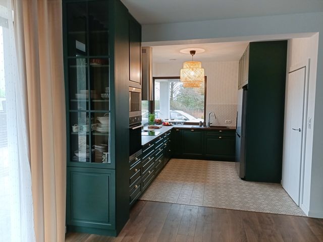 Nowoczesna, ciemnozielona kuchnia z drewnianą podłogą i wzorzystą posadzką w strefie roboczej, zrealizowana w Ulan-Majorat. Widoczna zabudowa kuchenna z witryną, liczne szuflady i sprzęt AGD.