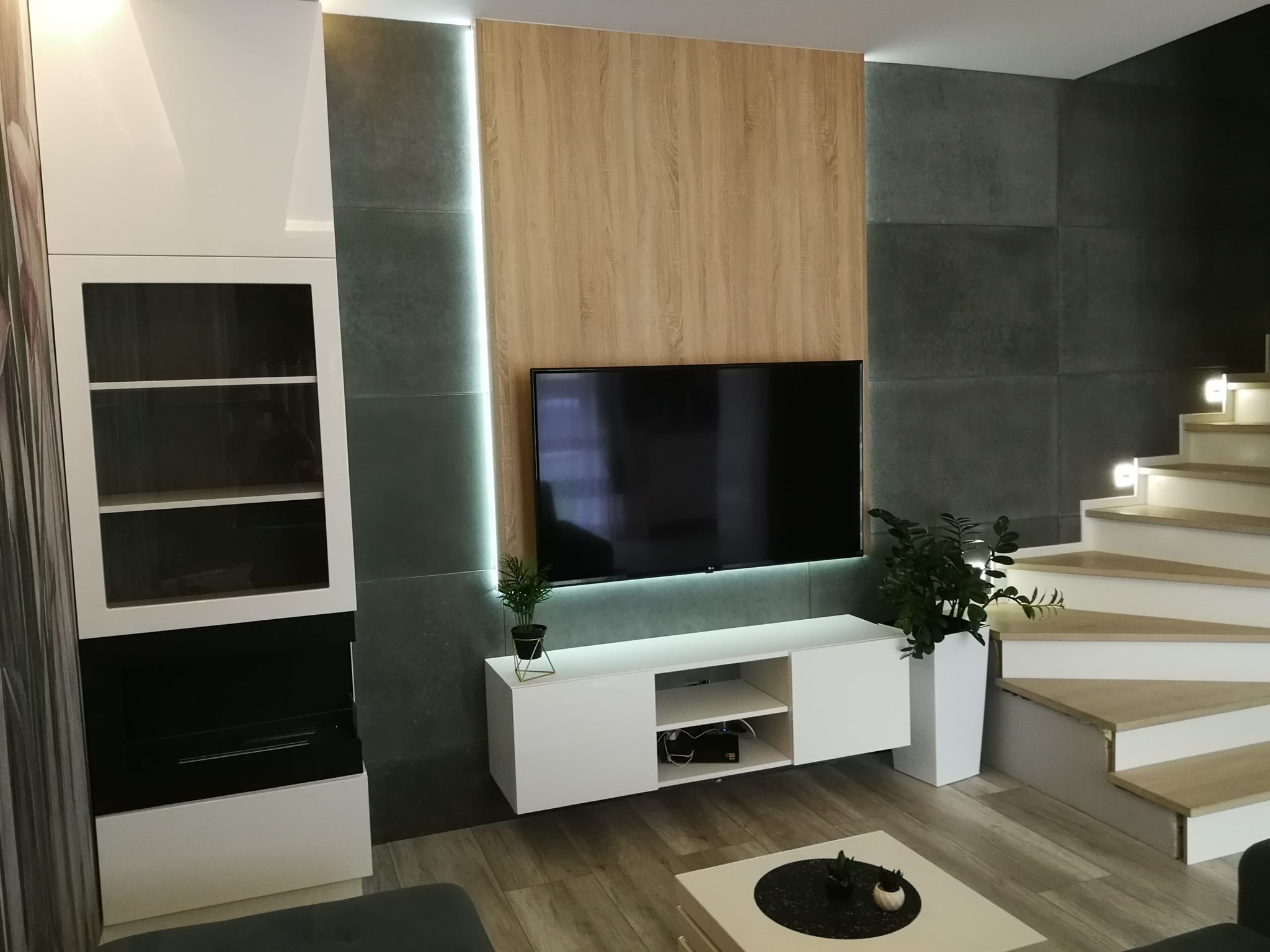 Nowoczesny salon z drewnianą ścianą telewizyjną podświetloną LED, białą szafką RTV, przeszkloną witryną i drewnianymi schodami z oświetleniem punktowym.