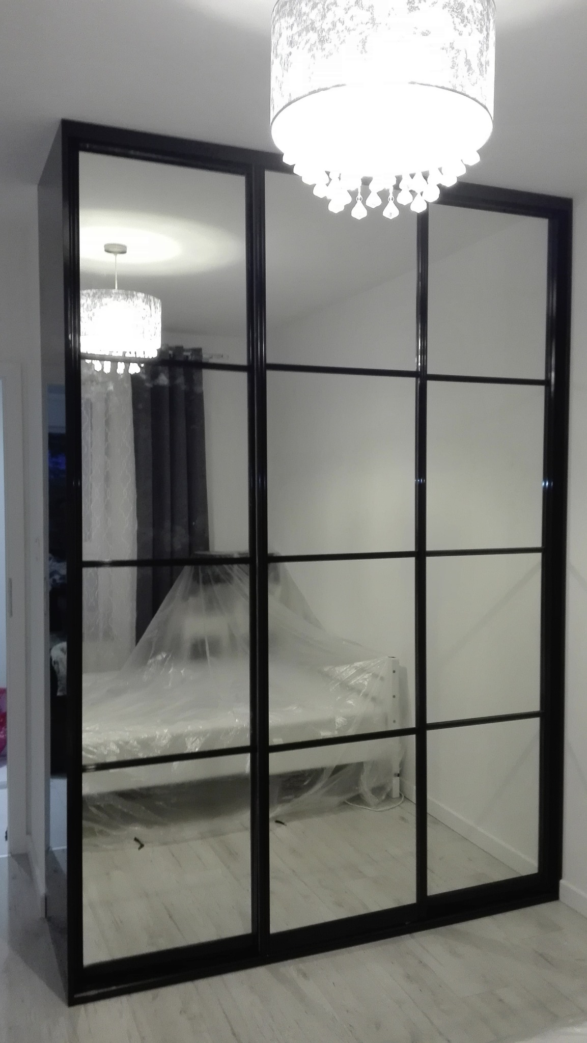 Czarna szafa przesuwna z lustrzanymi drzwiami, widok na łóżko przykryte folią ochronną i lampę sufitową