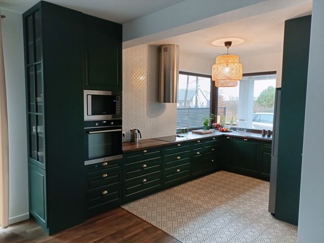 Nowoczesna kuchnia w odcieniach butelkowej zieleni z drewnianym blatem i wzorzystą podłogą, oświetlona plecioną lampą wiszącą, z widokiem na ogród za oknem.