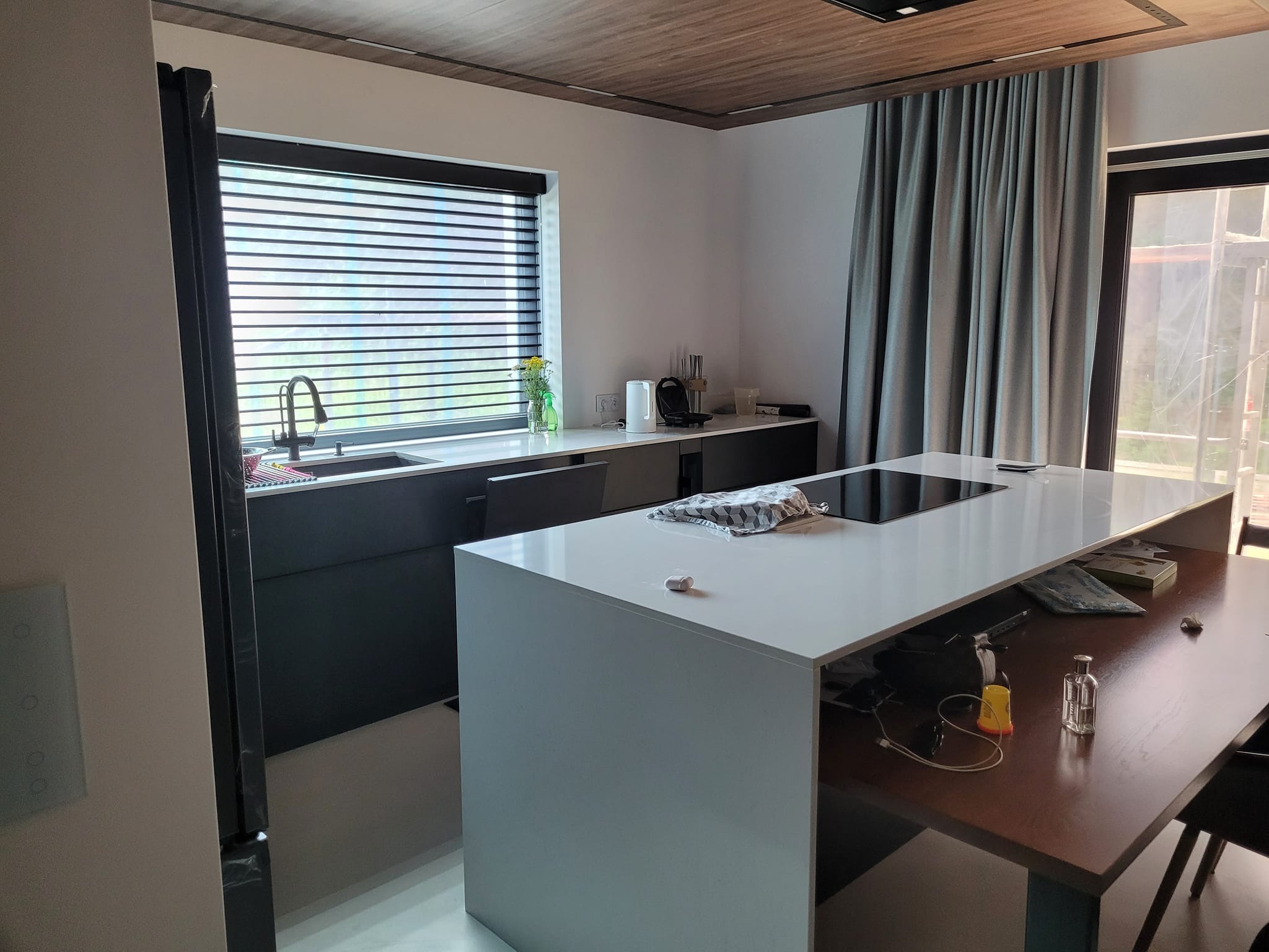 Nowoczesna kuchnia z wyspą w Ulan-Majorat, z czarnymi szafkami dolnymi i białym blatem, widok na okno z żaluzjami i drewniany sufit.