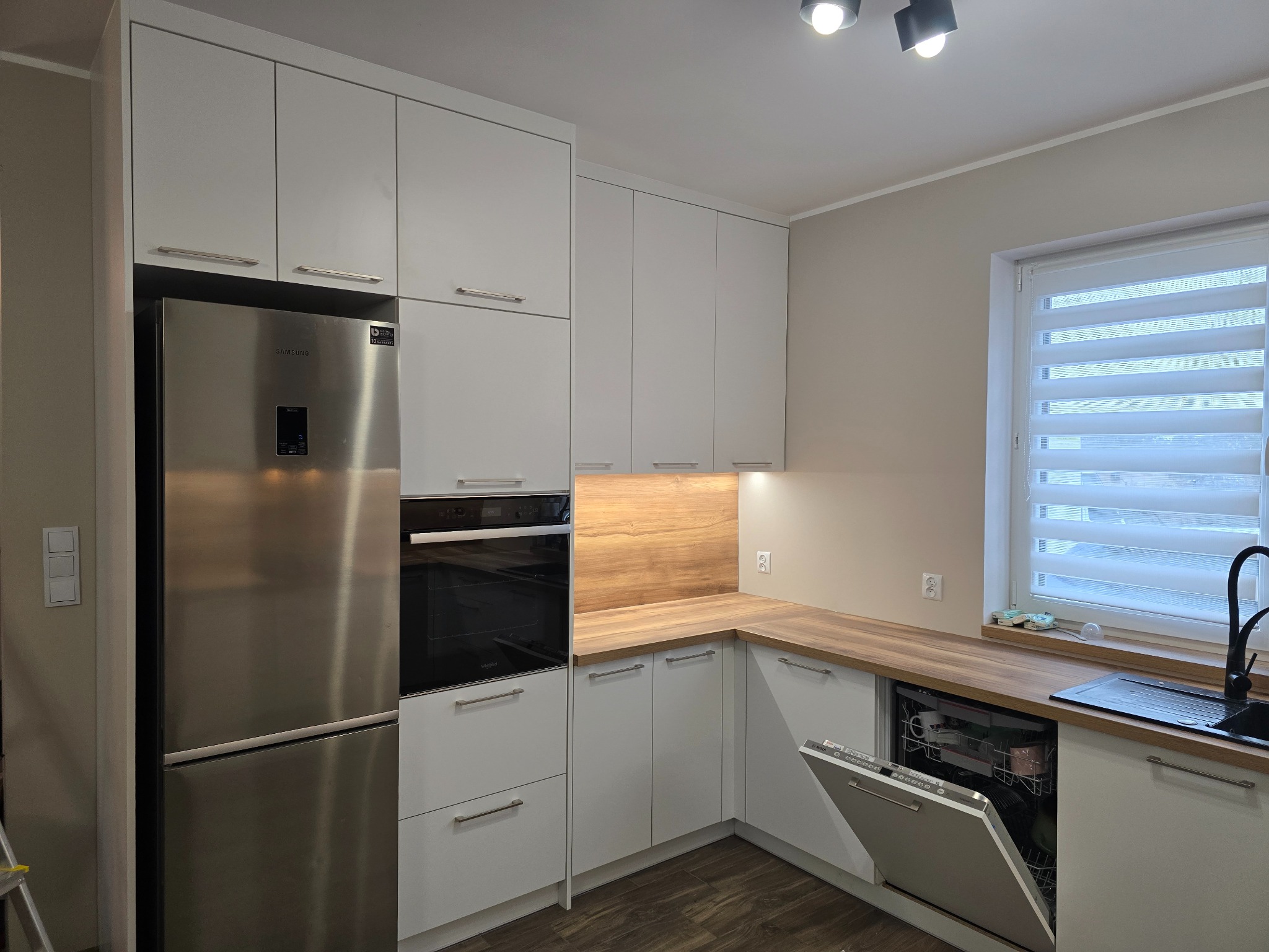 Nowoczesna kuchnia z białymi szafkami, drewnianym blatem, wbudowanym piekarnikiem i lodówką Samsung, z otwartą zmywarką.