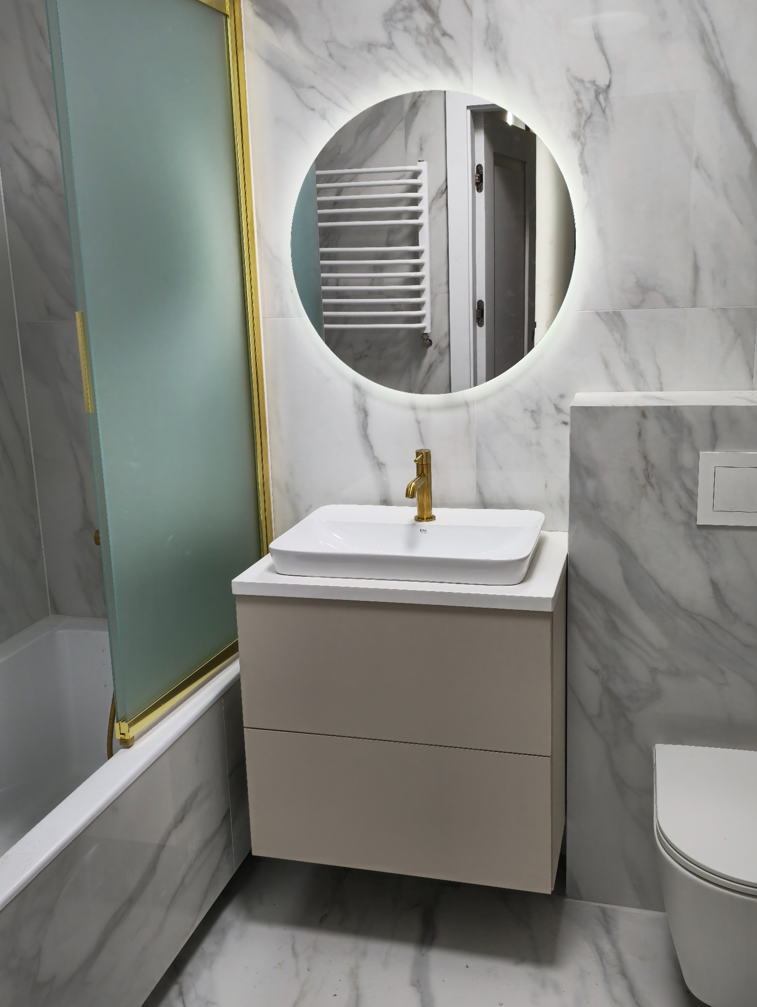 Nowoczesna łazienka z umywalką nablatową, szafką z szufladami w kolorze beżowym, baterią w kolorze złotym i okrągłym lustrem z podświetleniem LED na tle marmurowych płytek ściennych.