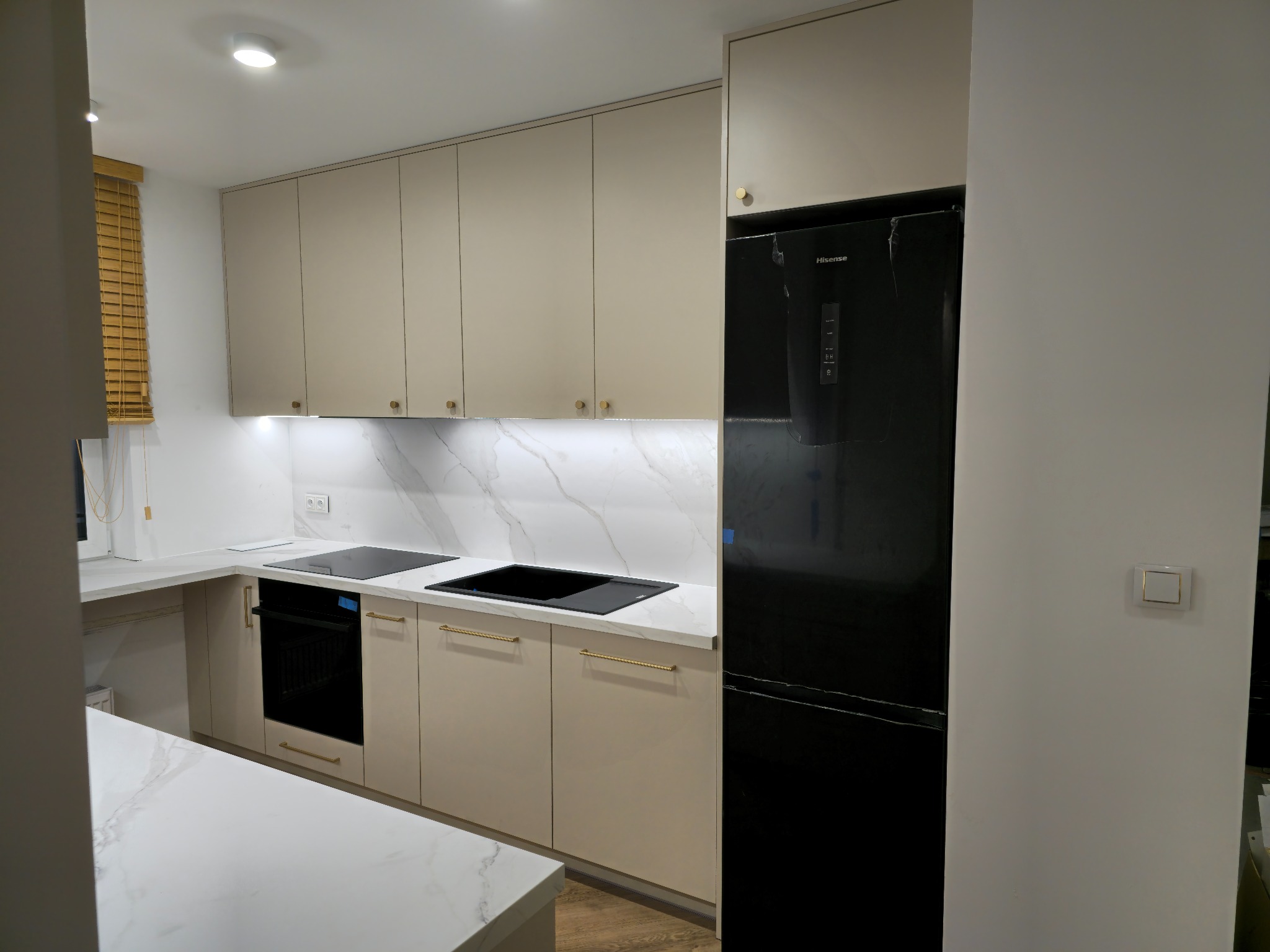 Nowoczesna kuchnia z beżowymi szafkami, białym blatem z marmurowym wzorem, czarną lodówką Hisense i złotymi uchwytami.