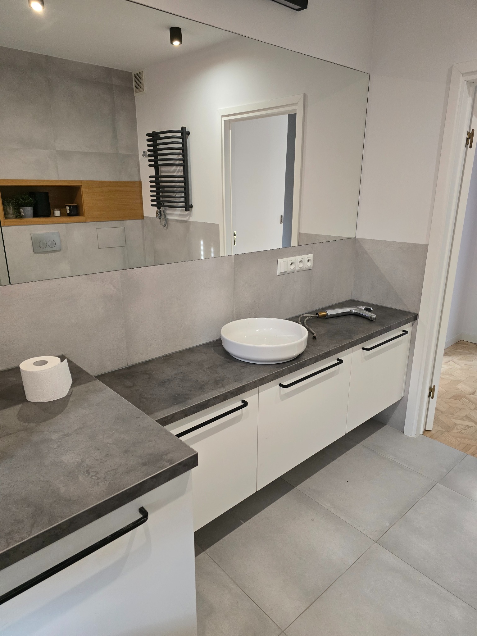 Nowoczesna łazienka z białymi szafkami pod umywalką, szarym blatem i płytkami. Widoczne czarne uchwyty i grzejnik. Lustro odbija fragment wnętrza.