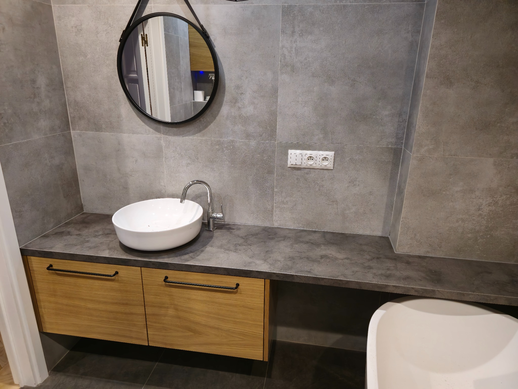Nowoczesna łazienka z okrągłym lustrem i umywalką nablatową na szafce z drewnianymi frontami, szare płytki na ścianach i podłodze, widoczne gniazdka elektryczne.
