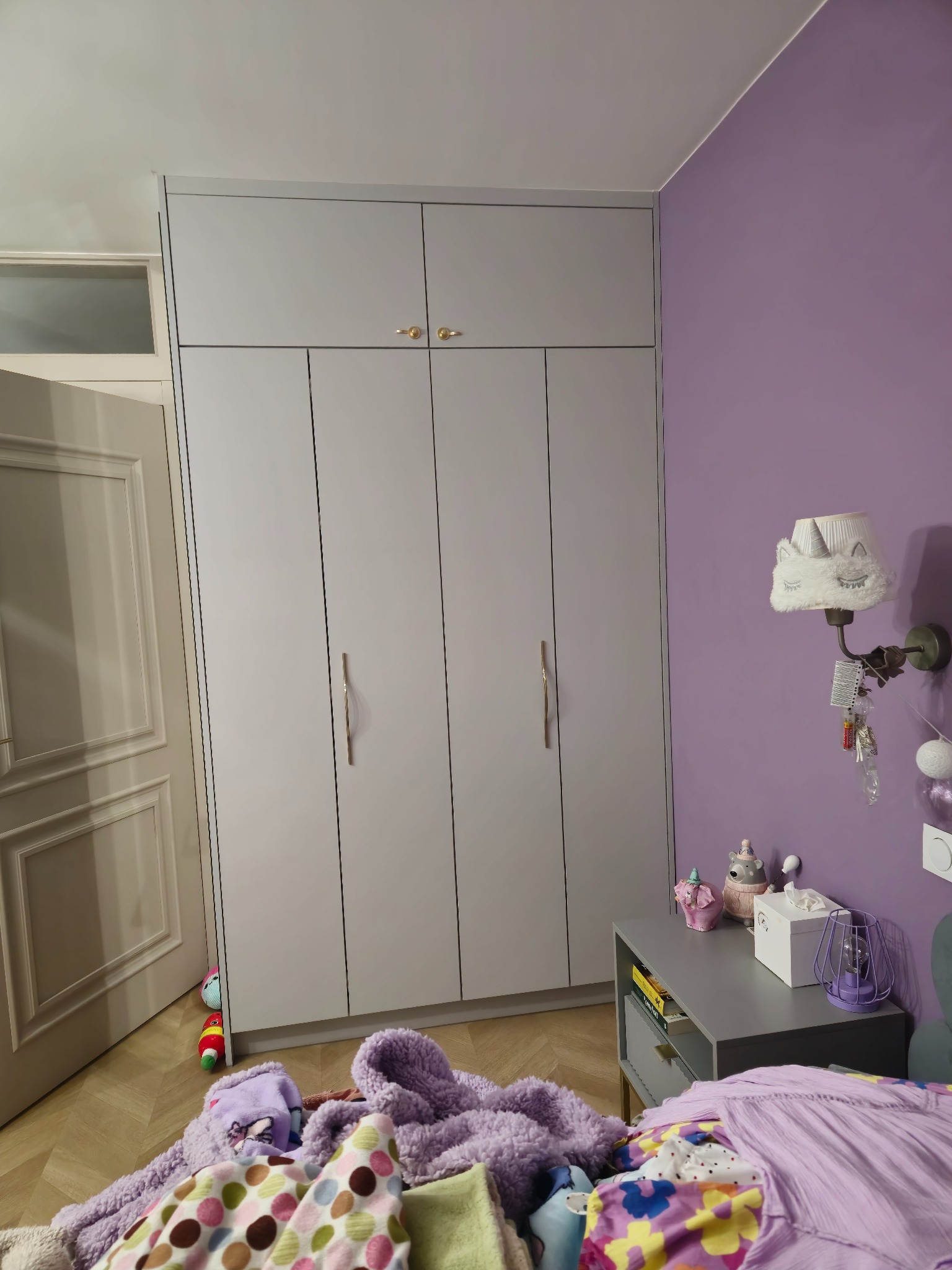 Szara szafa na wymiar z złotymi uchwytami w pokoju dziecka z fioletowymi ścianami. Na podłodze leżą zabawki i ubrania, co dodaje wnętrzu przytulności.