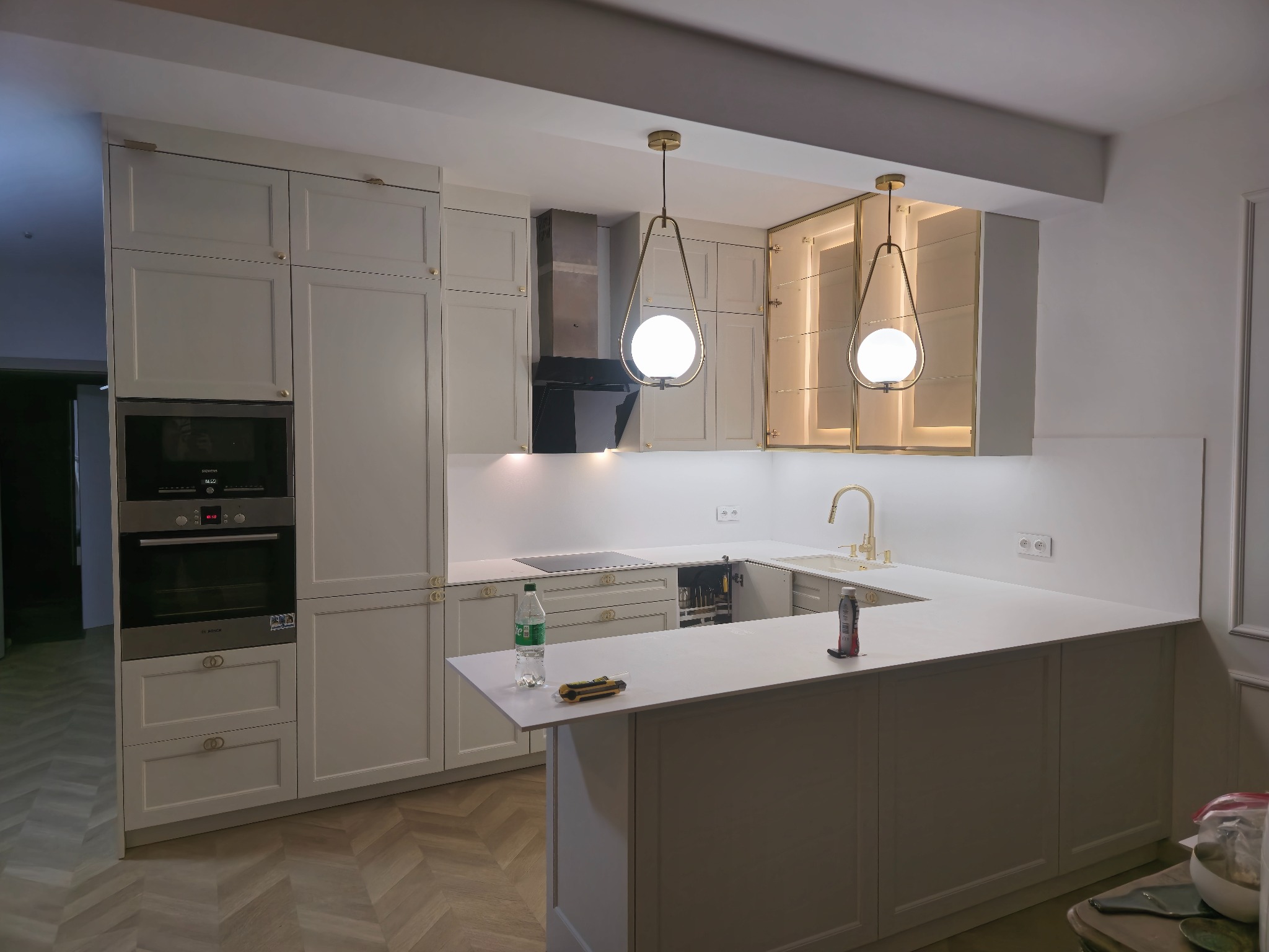 Jasna kuchnia z frezowanymi frontami, złotymi uchwytami i lampami. Na blacie leżą narzędzia, co sugeruje świeży montaż. Parkiet ułożony w jodełkę.