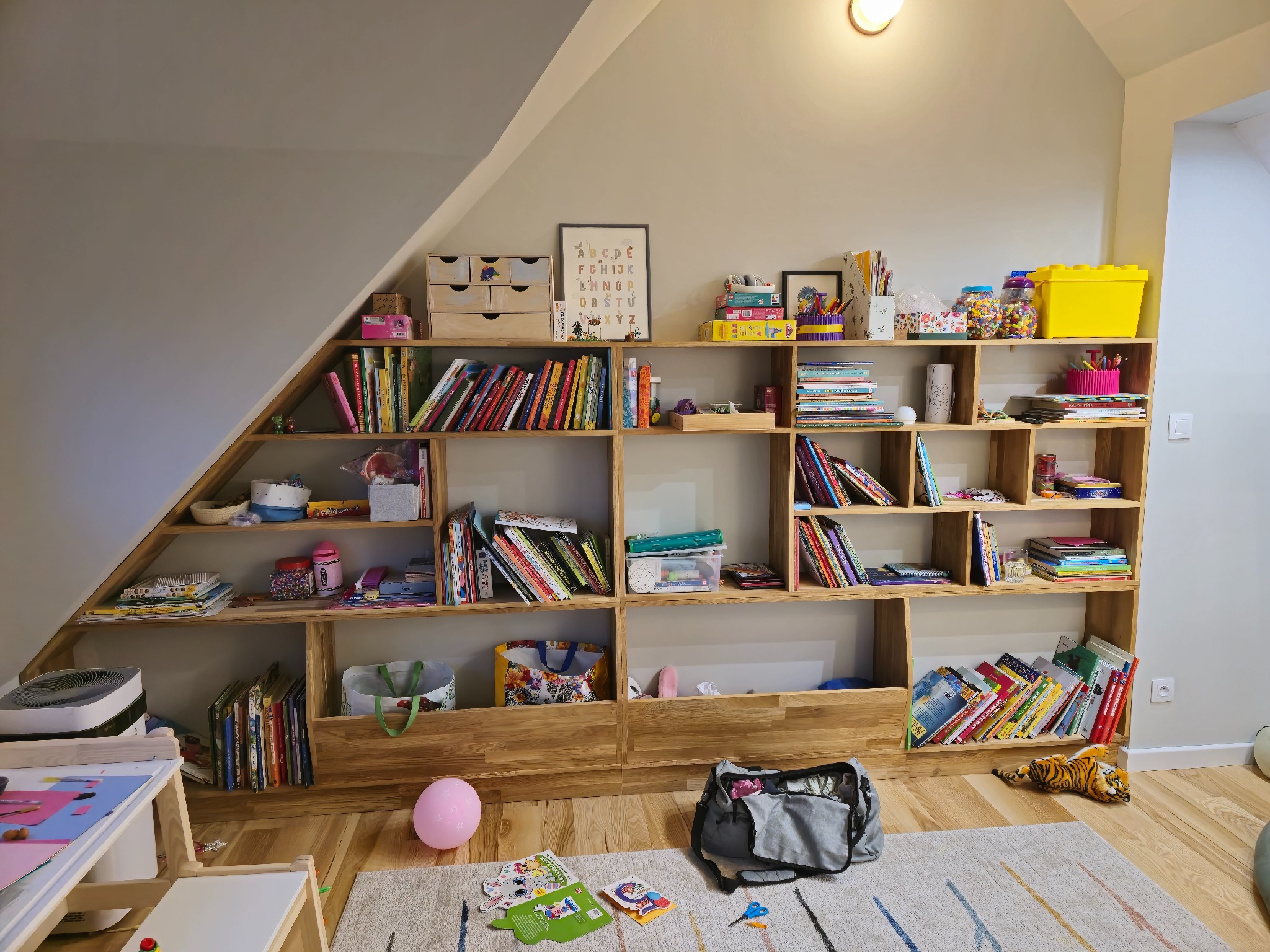 Drewniana, nieregularna biblioteczka pod skosem na poddaszu, wypełniona książkami, zabawkami i dekoracjami w pokoju dziecka. Widoczny fragment dywanu i biurka.