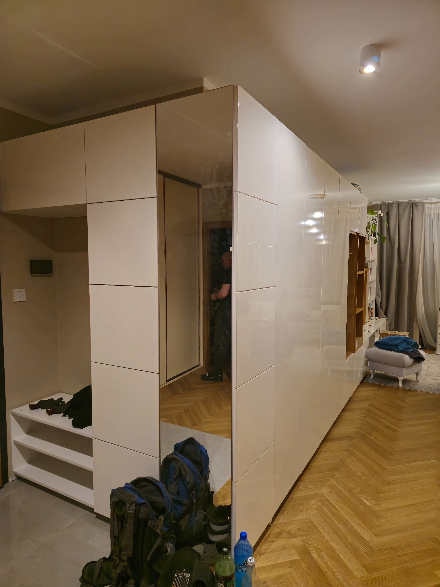 Biała, lakierowana szafa na wymiar z lustrzanymi drzwiami przesuwnymi, tworząca garderobę i przedpokój w nowoczesnym mieszkaniu z parkietem w jodełkę.