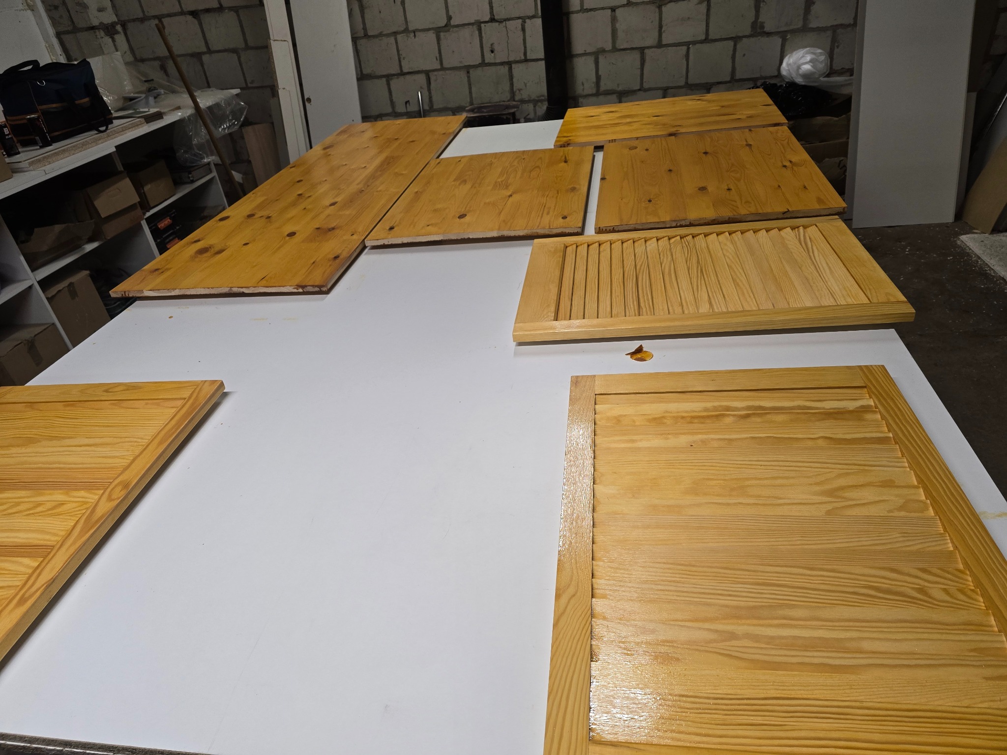 Lakierowane, drewniane fronty meblowe (w tym żaluzjowe) leżą na białym stole w warsztacie. Widoczne słoje drewna i połysk lakieru. Część elementów poza kadrem.