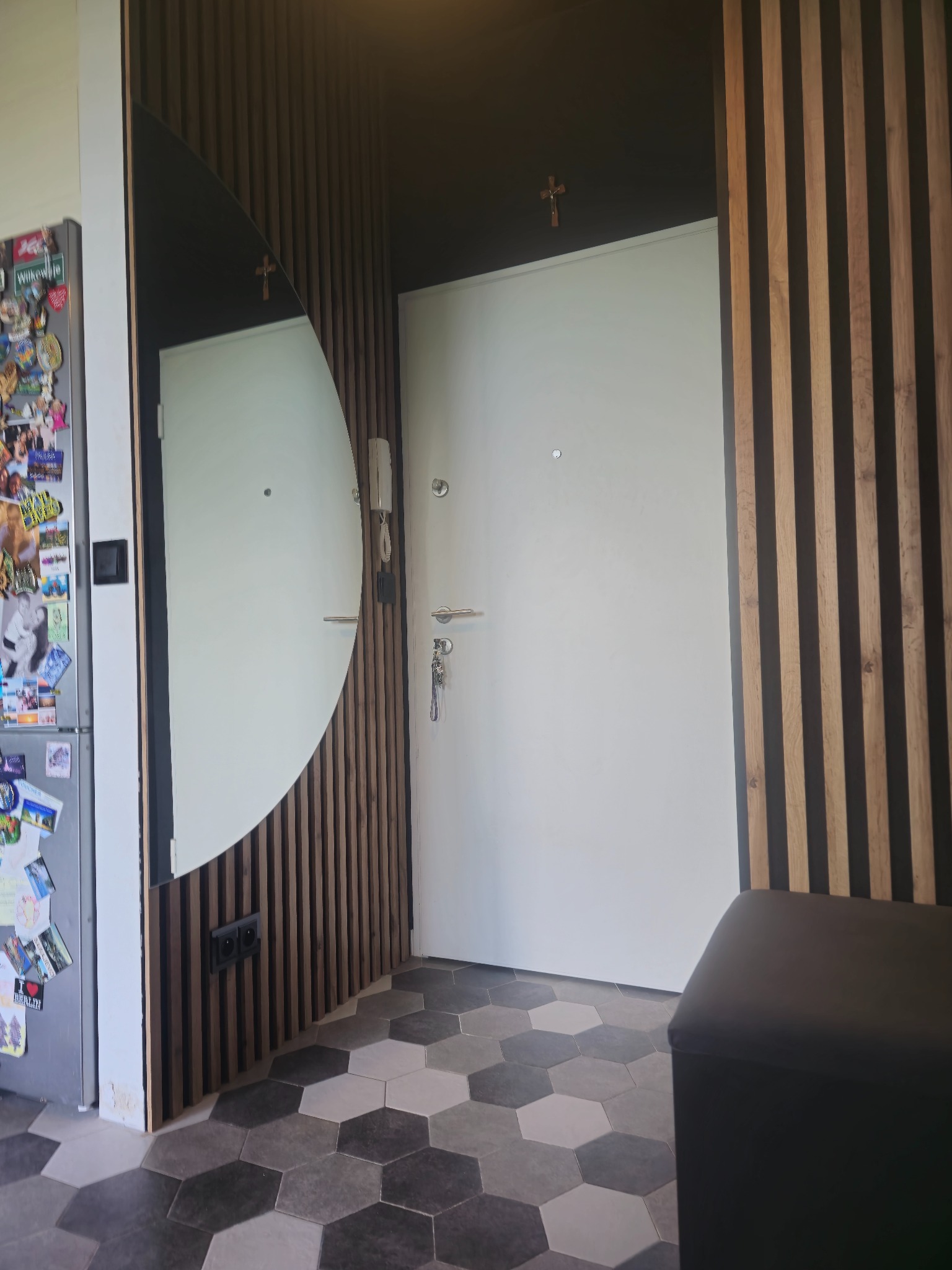 Nowoczesna aranżacja przedpokoju z lamelami drewnianymi, owalnym lustrem i heksagonalną podłogą. Nad drzwiami widoczny krzyż. Lodówka z magnesami po lewej.