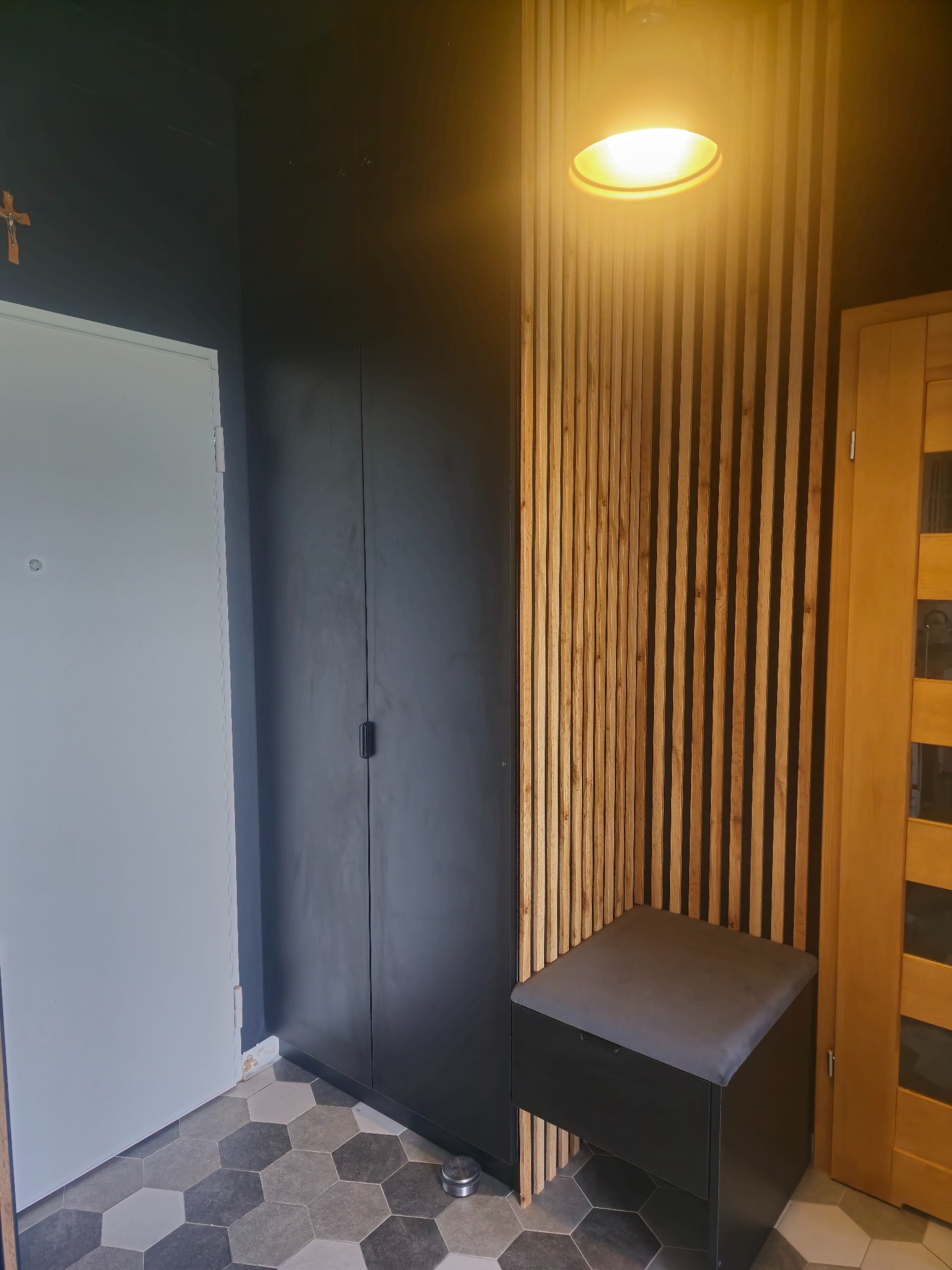 Nowoczesny przedpokój: szara szafa w zabudowie, drewniana ażurowa ścianka, siedzisko i heksagonalna podłoga. Minimalistyczny design z akcentem naturalnego drewna.