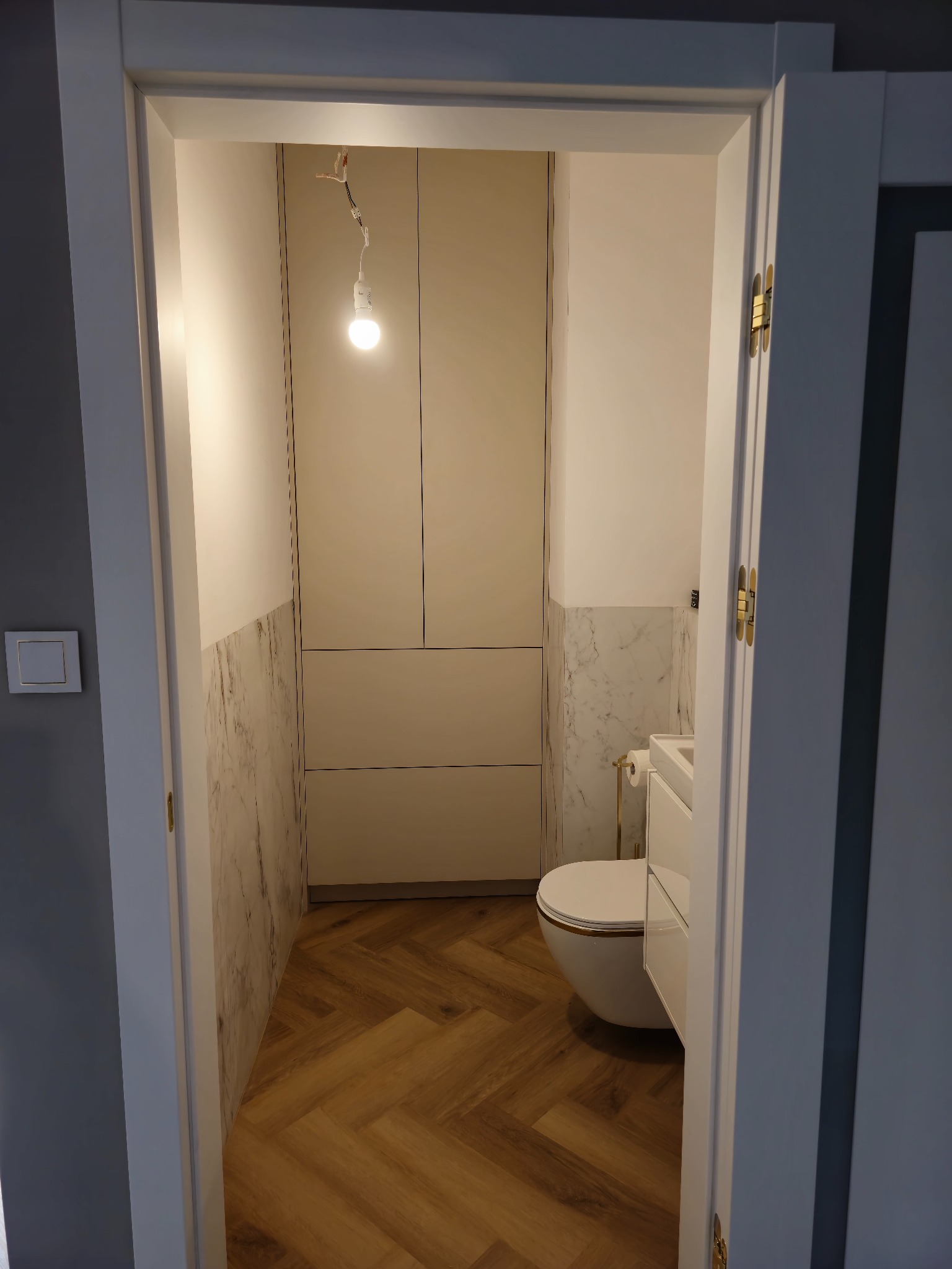 Nowoczesna szafa wnękowa w łazience w Ulan-Majorat. Białe fronty, marmurowe wykończenie ścian, parkiet w jodełkę. Widok z korytarza przez otwarte drzwi.