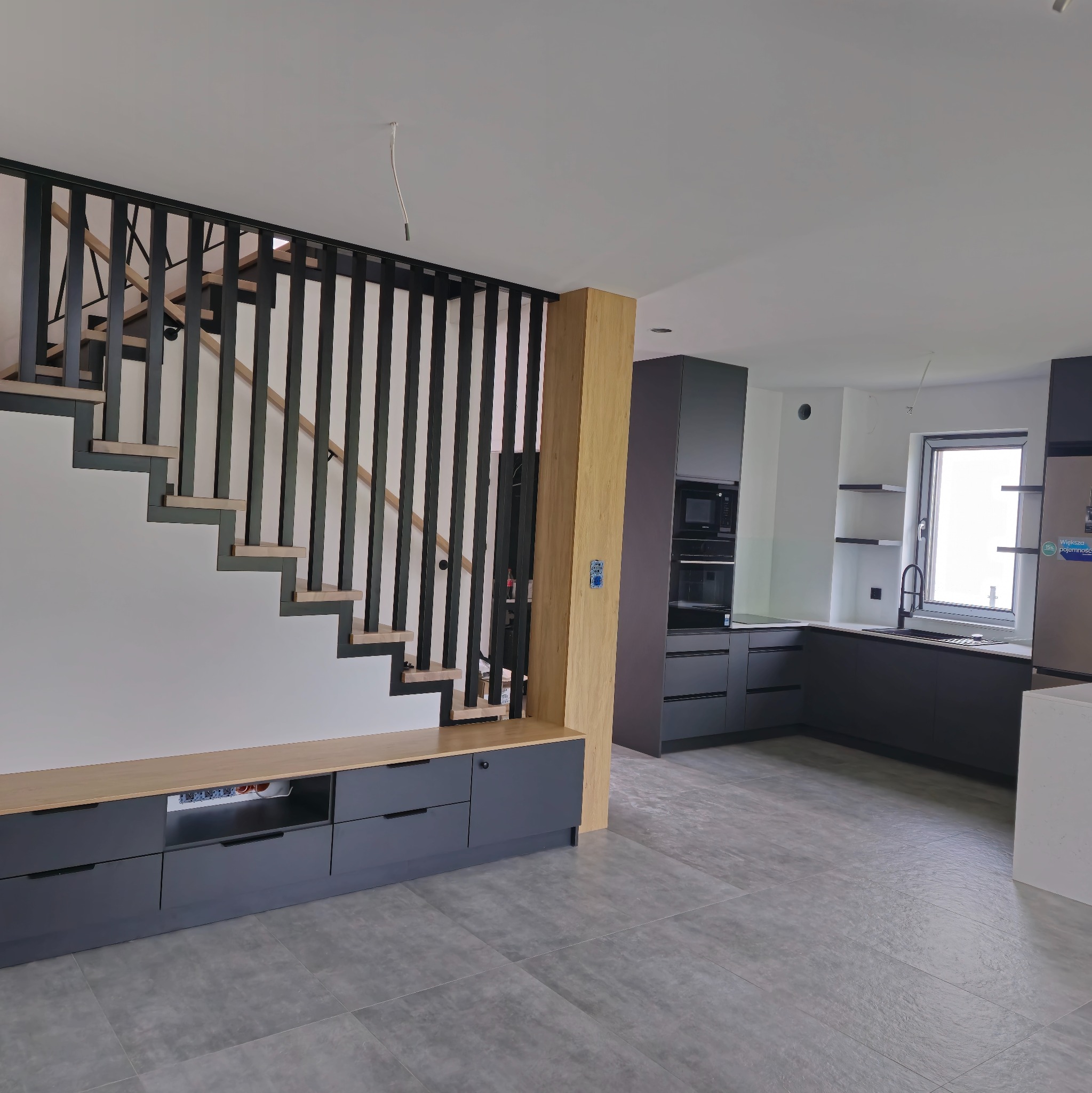 Nowoczesna kuchnia z szarymi meblami i drewnianymi akcentami, schody z czarną balustradą, szara podłoga. Minimalistyczny design.