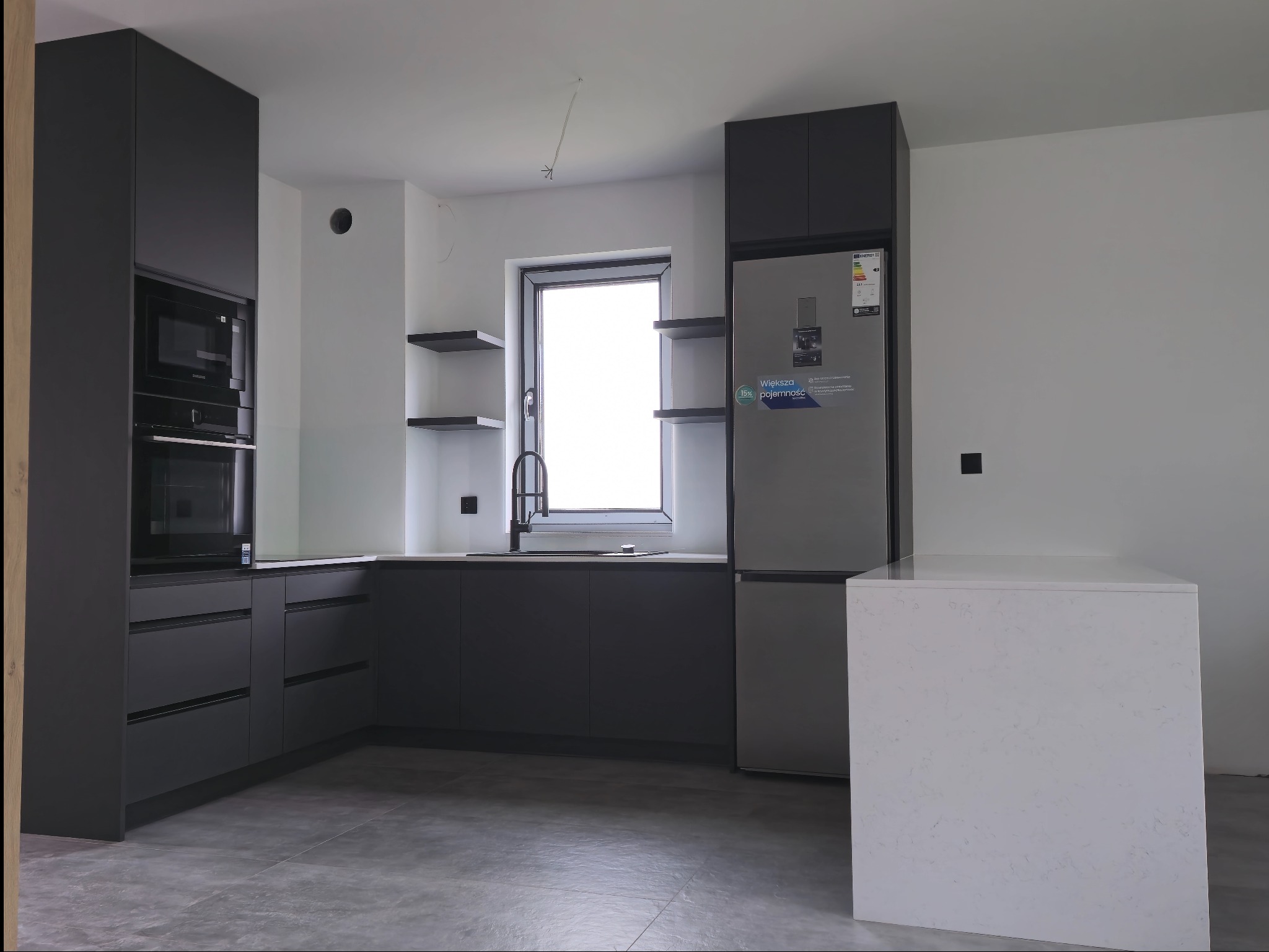 Nowoczesna, minimalistyczna kuchnia z grafitowymi frontami, wyspą z białym blatem i sprzętem AGD w zabudowie. Widoczne półki wiszące i szara podłoga.
