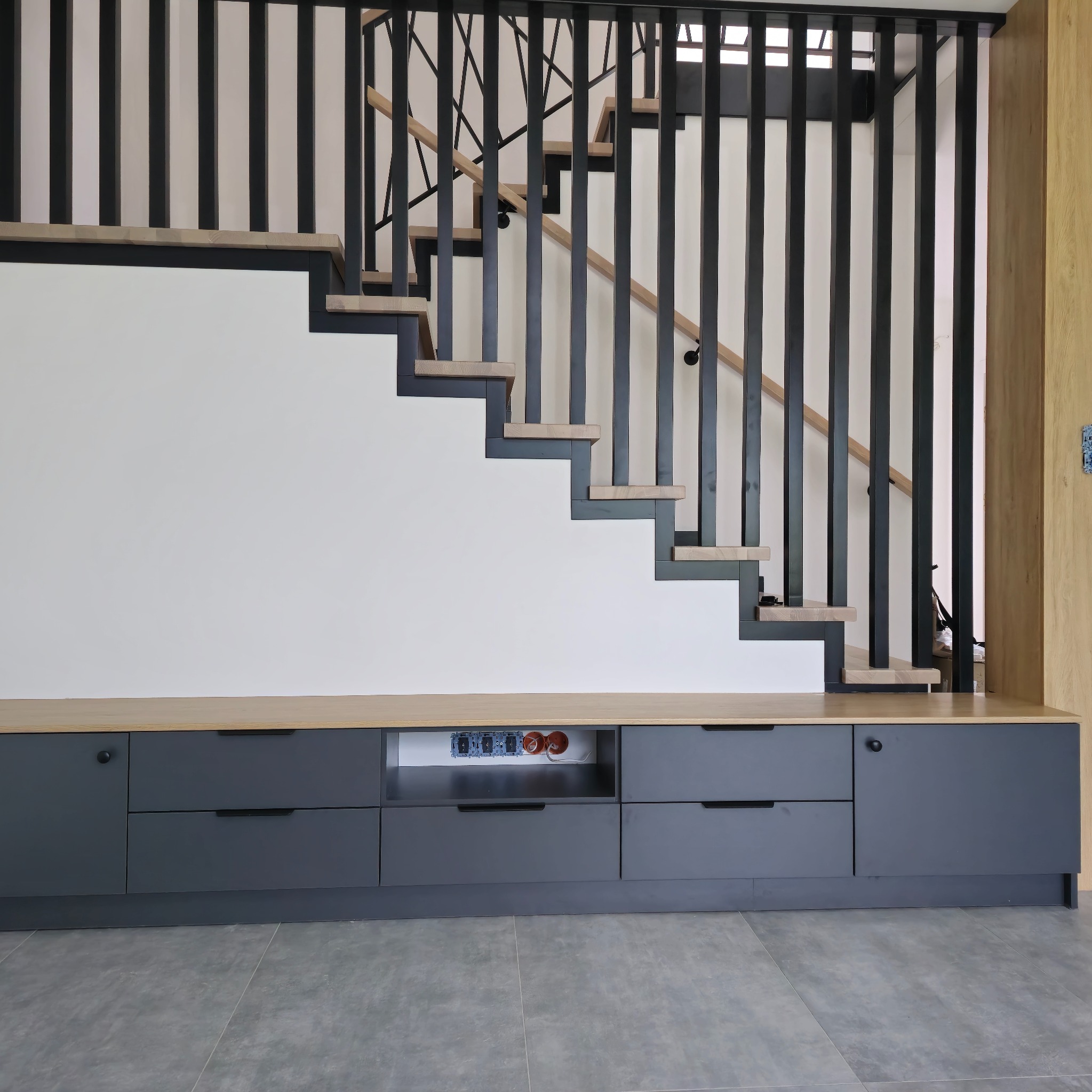 Nowoczesne schody z czarną balustradą i drewnianymi stopniami, zintegrowane z szarą szafką na ścianie. Widoczne gniazdka elektryczne. Minimalistyczny design.