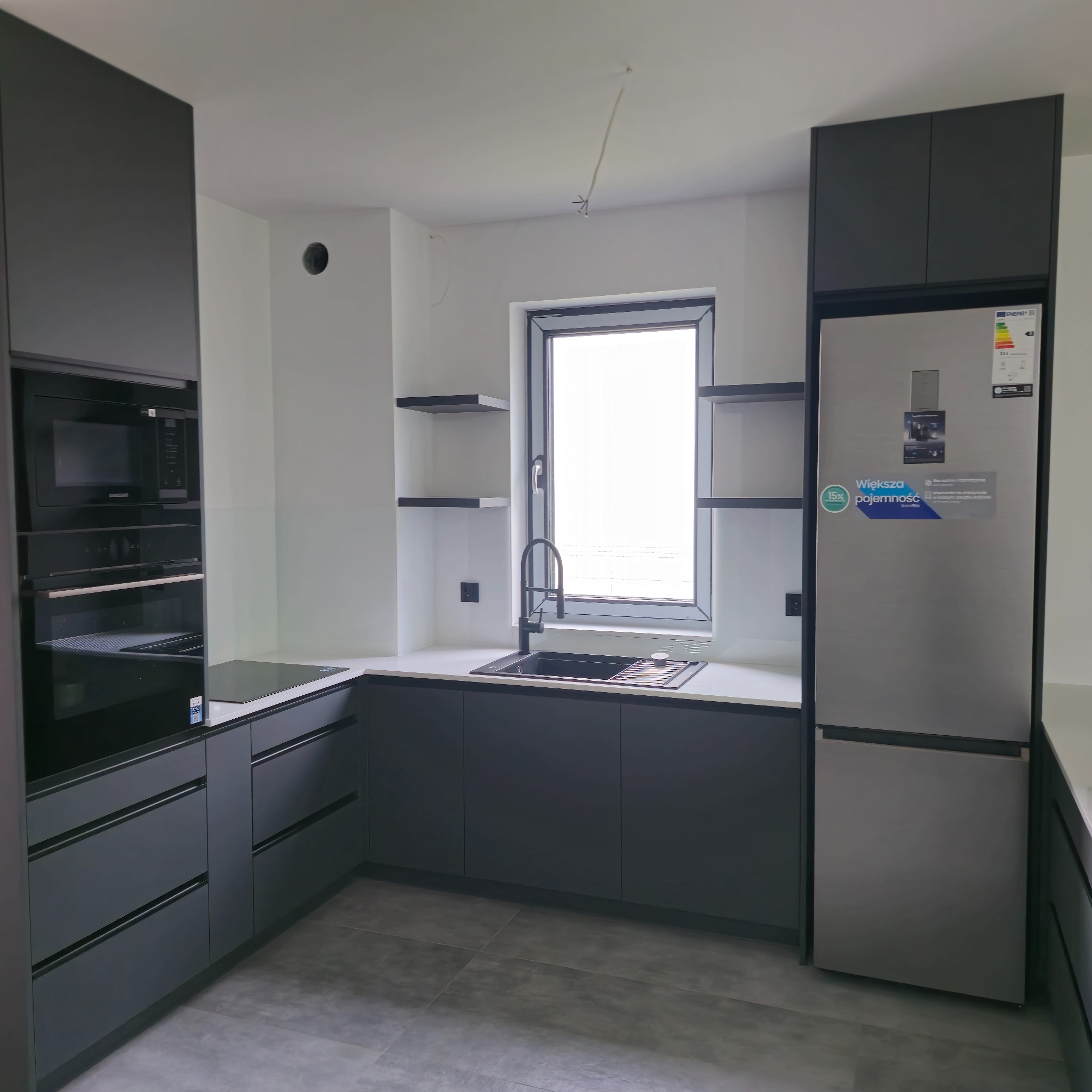 Nowoczesna kuchnia w odcieniach szarości z białym blatem i srebrną lodówką. Minimalistyczny design z otwartymi półkami i zabudowanym sprzętem AGD.