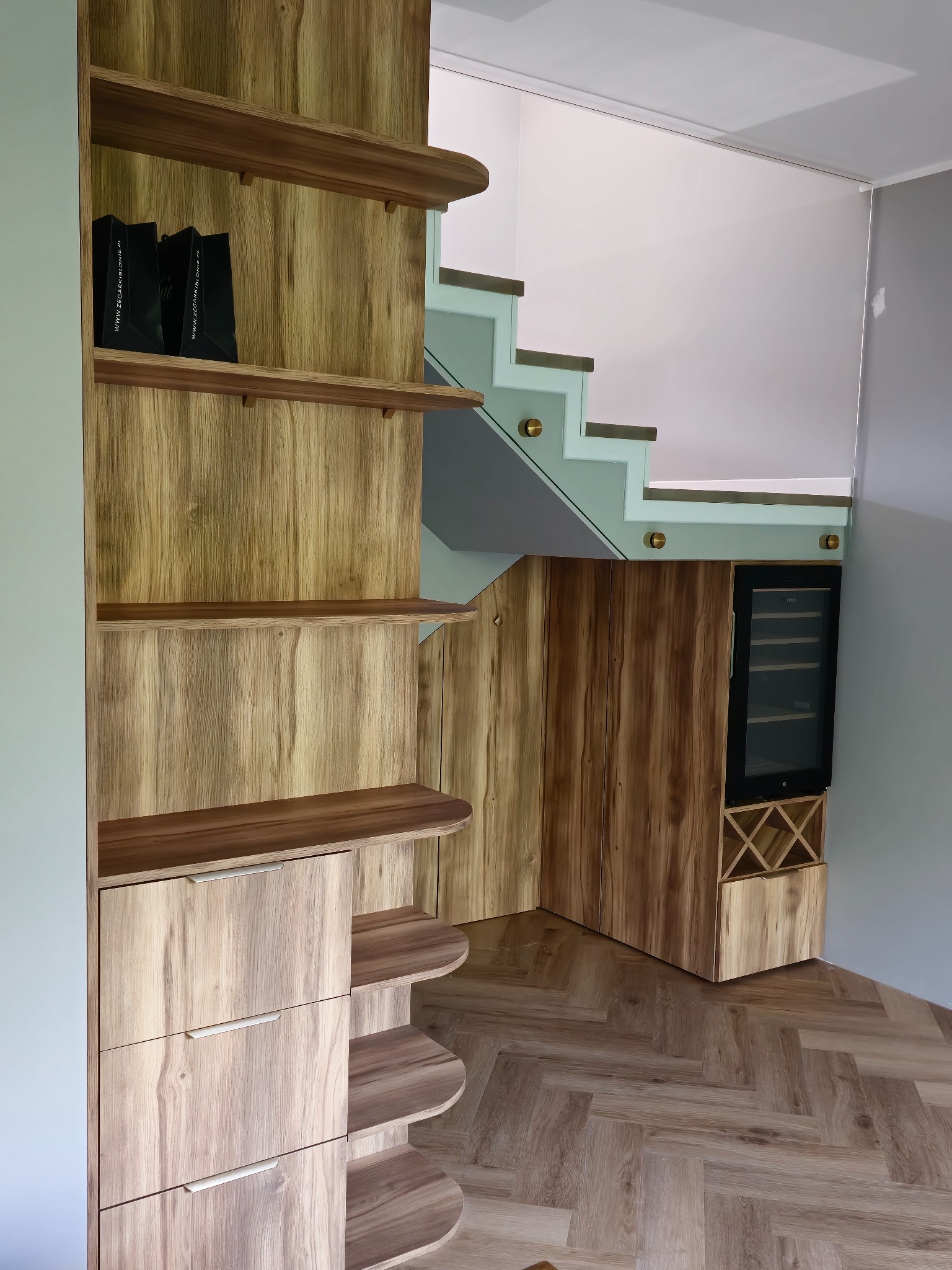 Drewniana zabudowa pod schodami z półkami, szafkami i chłodziarką do wina. Parkiet ułożony w jodełkę. Nowoczesne schody ze szklaną balustradą.