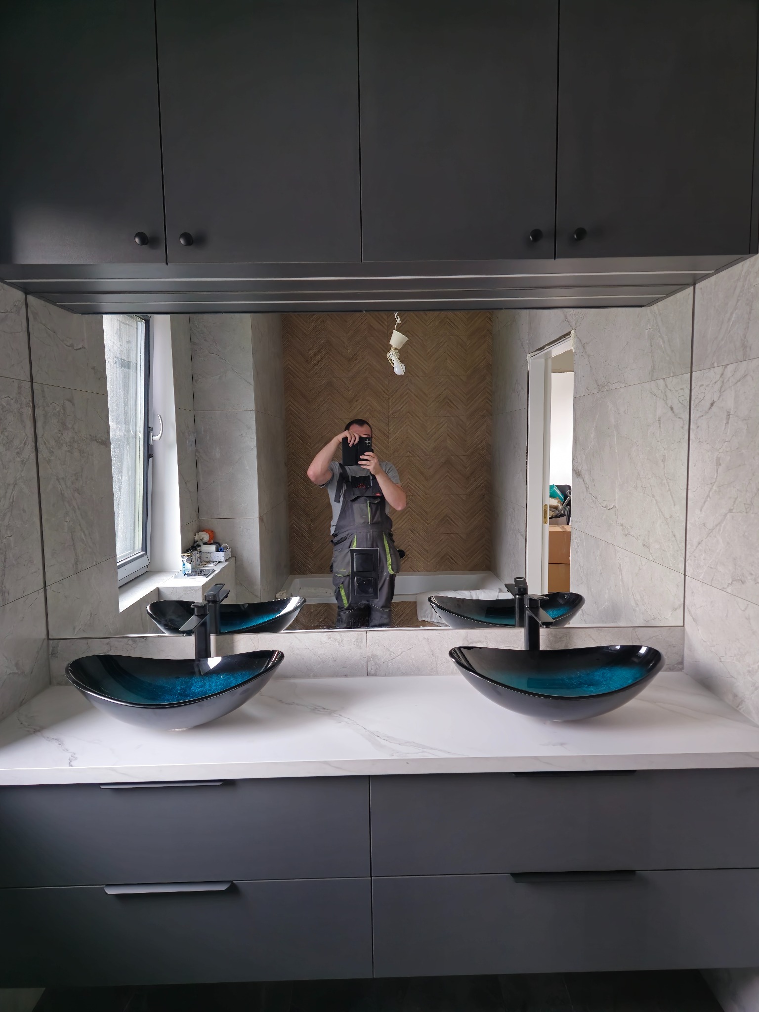 Nowoczesna łazienka z szarą zabudową meblową, umywalkami w kształcie mis i dużym lustrem. Widoczne płytki imitujące marmur i oświetlenie dekoracyjne.